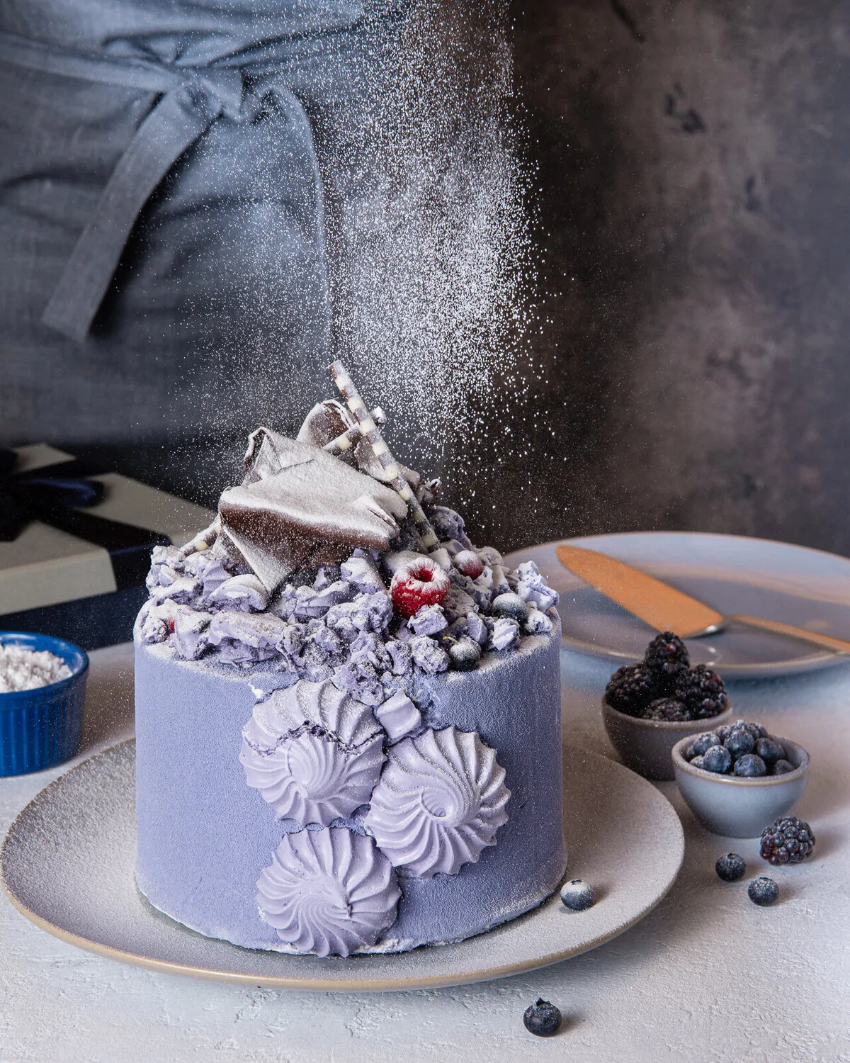 Icing sugar falling over a purple cake decorated with berries and chocolate shards.
