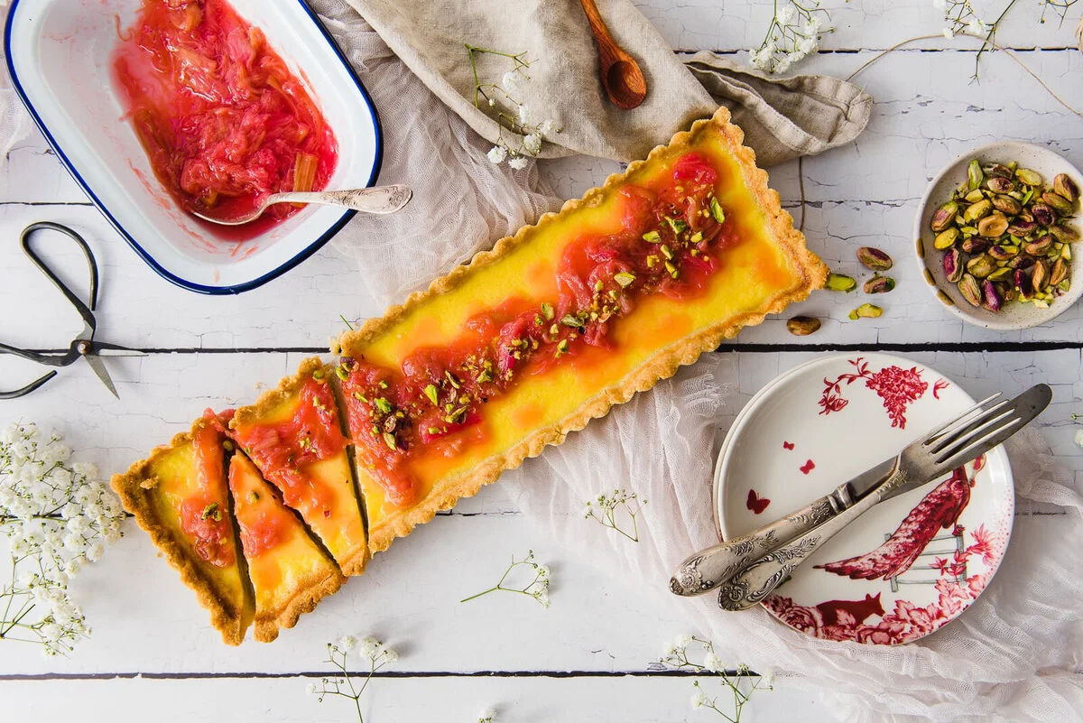 Long rhubarb and labneh tart on a pale table with serving utensils.