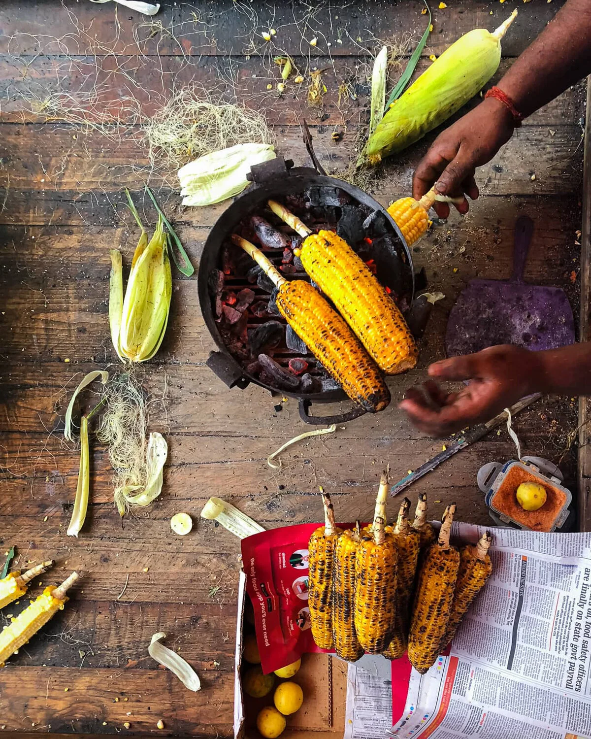 Hands seasoning charred corn beside a pan of corn cobs.