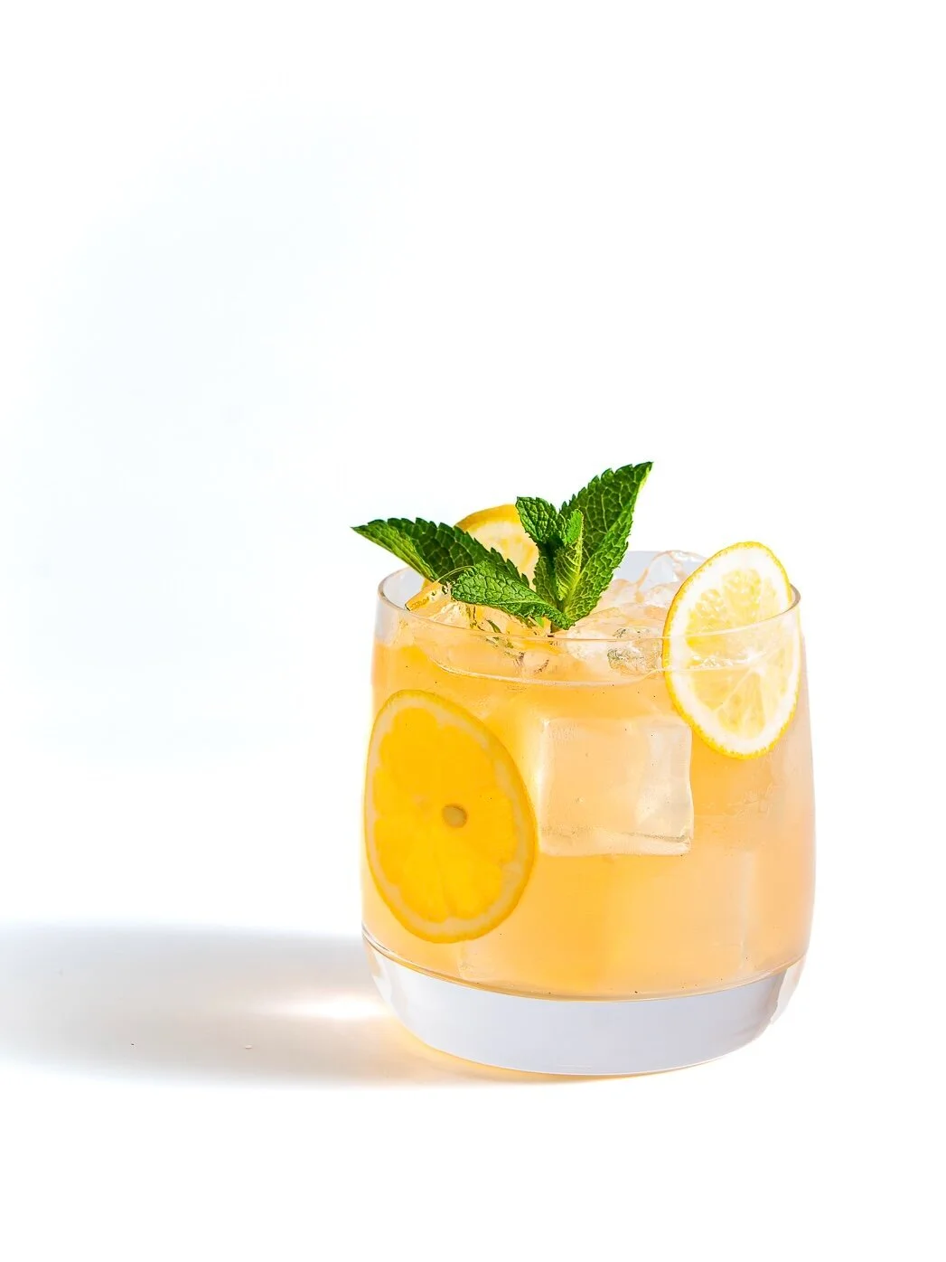 Pale yellow drink with ice, citrus and mint against a white backdrop.