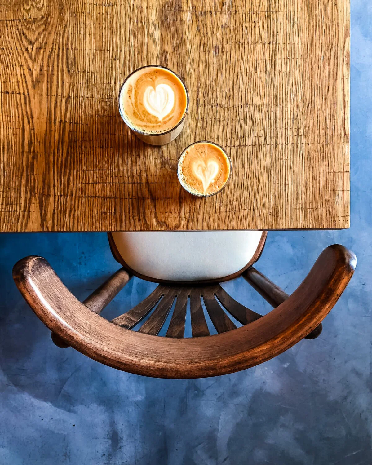Overhead view of two coffees on a wooden table beside a blue chair.